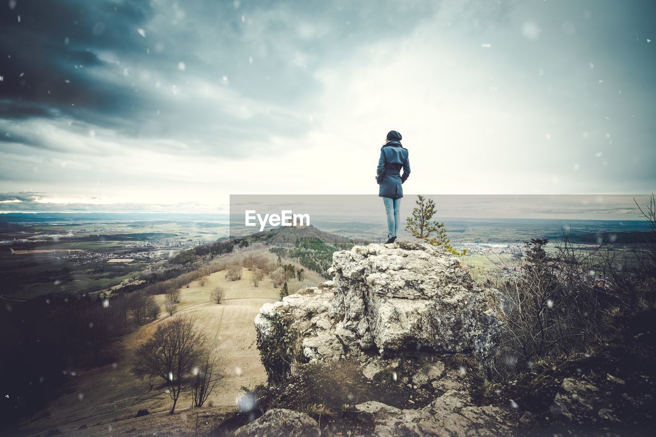 Rear view of woman standing on cliff against cloudy sky during snowfall