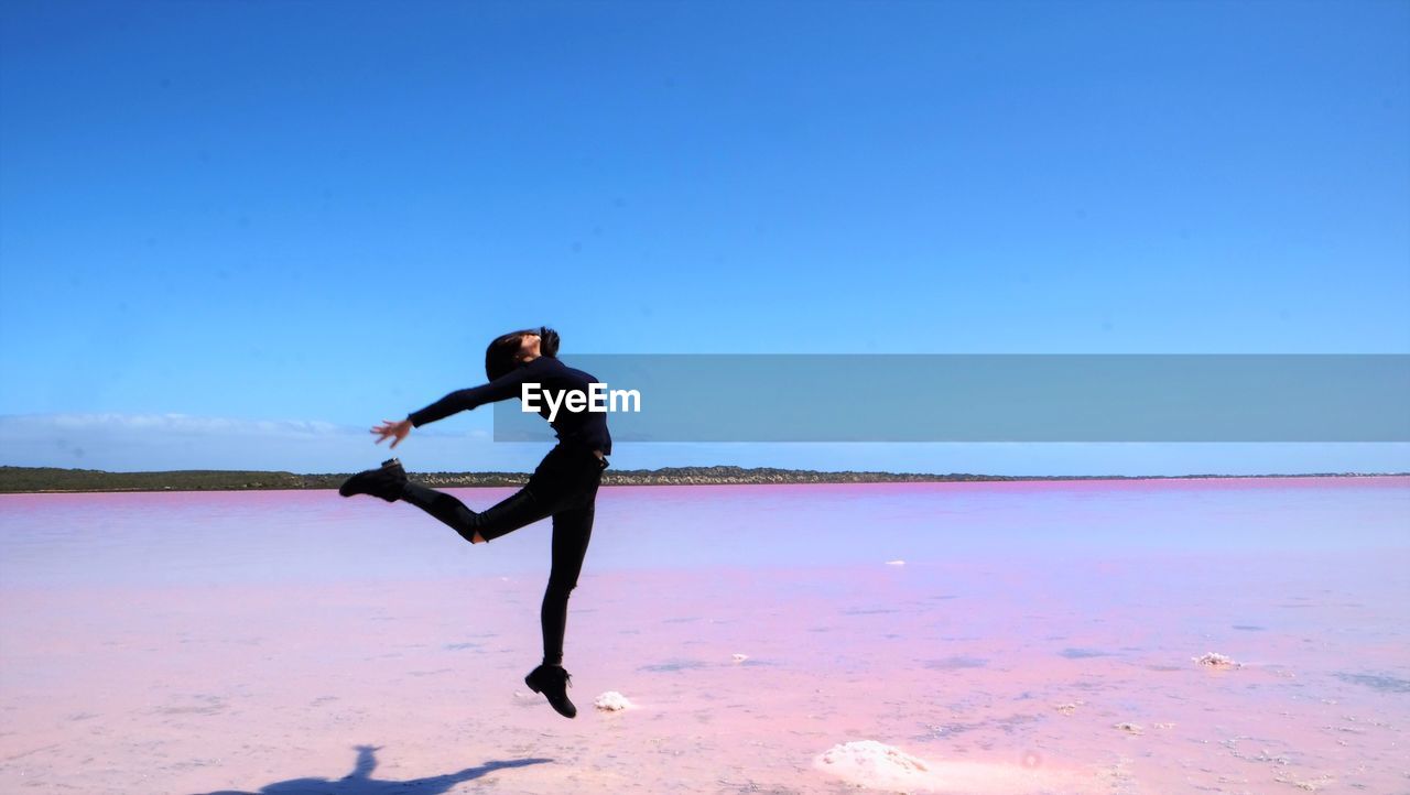 Side view of woman jumping over pink lake against blue sky