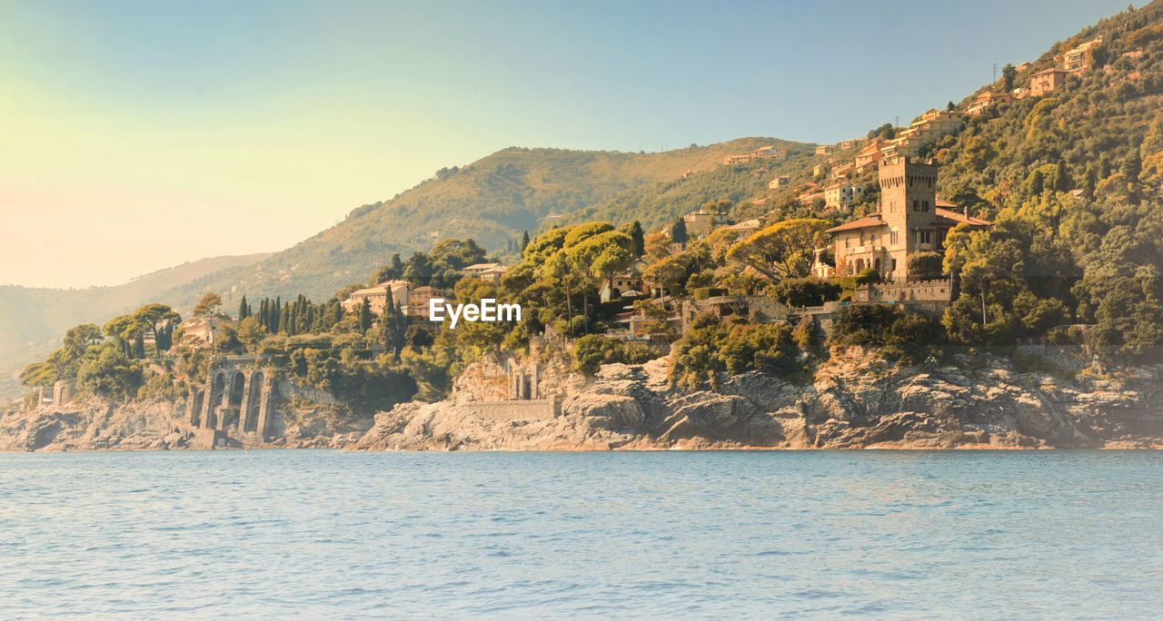 Rippled river water with houses and trees on cliff against clear sky