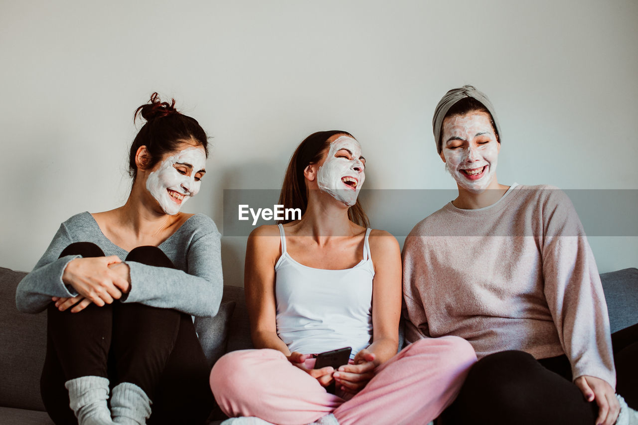 Cheerful female friends with facial masks sitting against wall at home