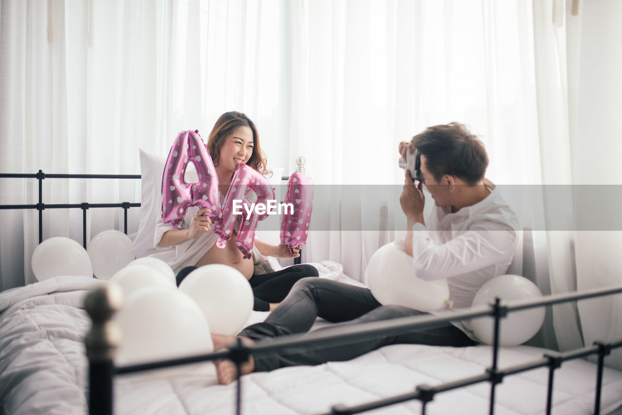 Man photographing pregnant wife holding props at bedroom