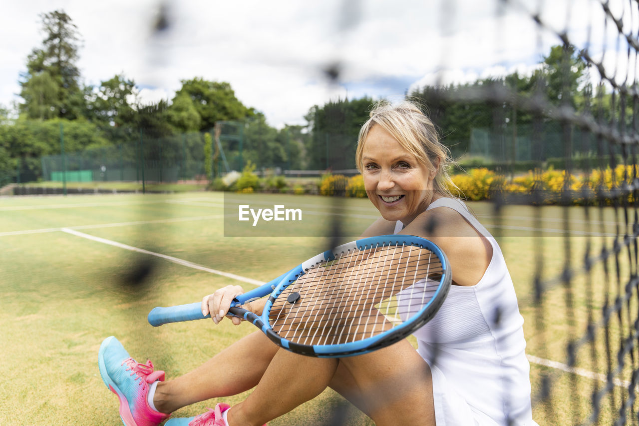 Portrait of smiling mature woman sitting on grass court at tennis club