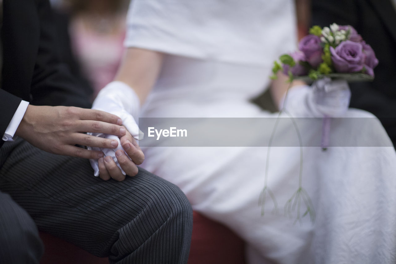 Midsection of bride and groom holding hands and flower bouquet