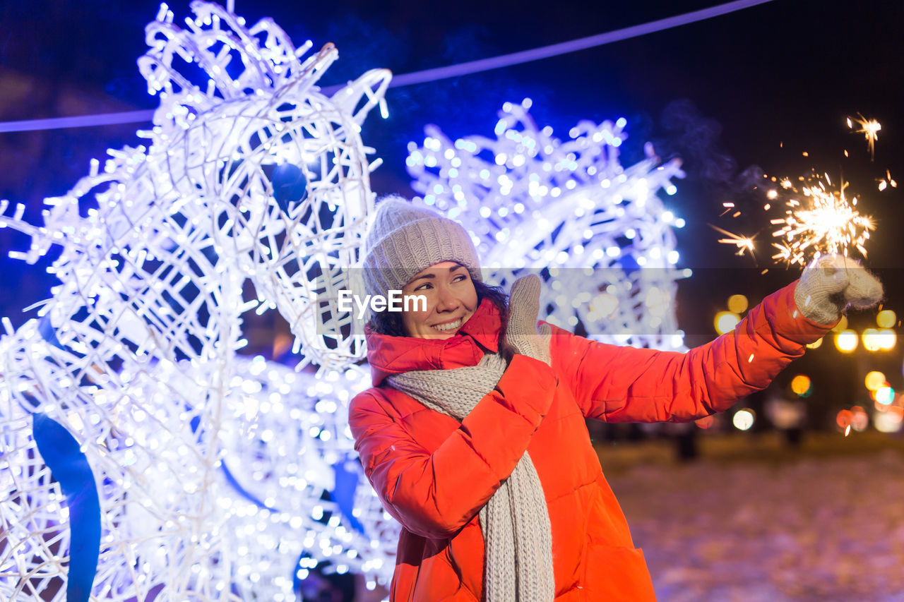 portrait of smiling woman with arms raised standing against illuminated christmas tree