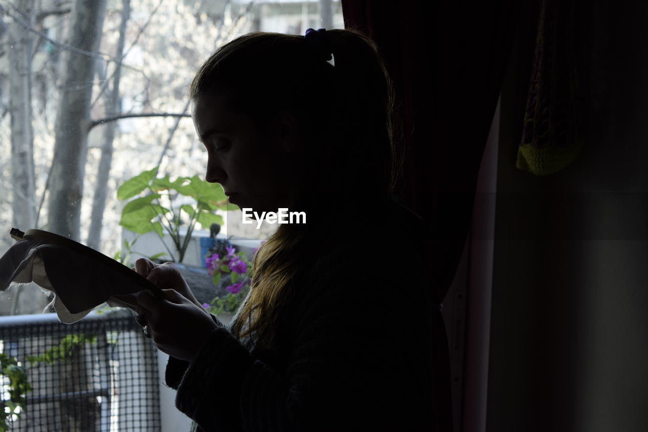 Side view of young woman embroidering at window