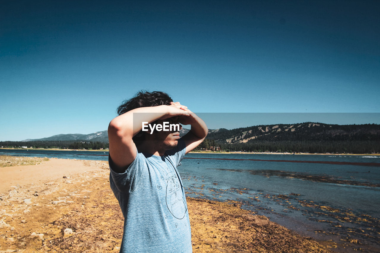 Man shielding eyes while standing at lakeshore against clear sky