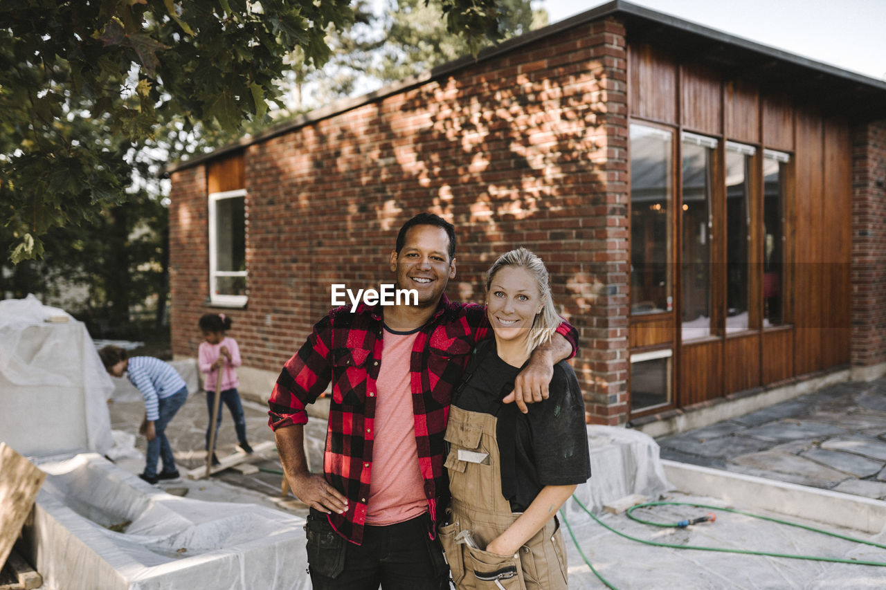 Portrait of confident couple standing while children playing in background against house