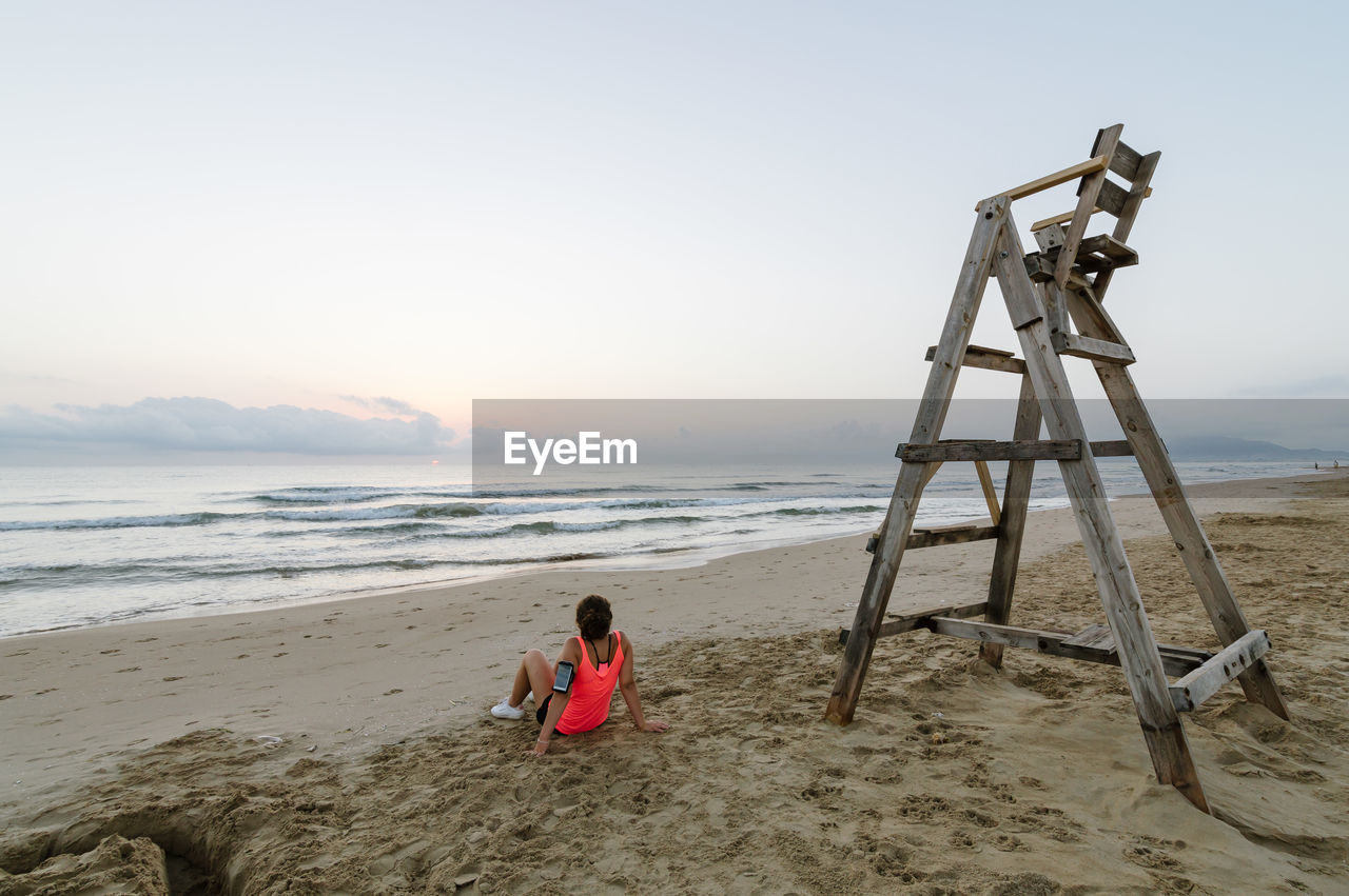 Rear view of woman sitting by wooden ladder at beach against sky