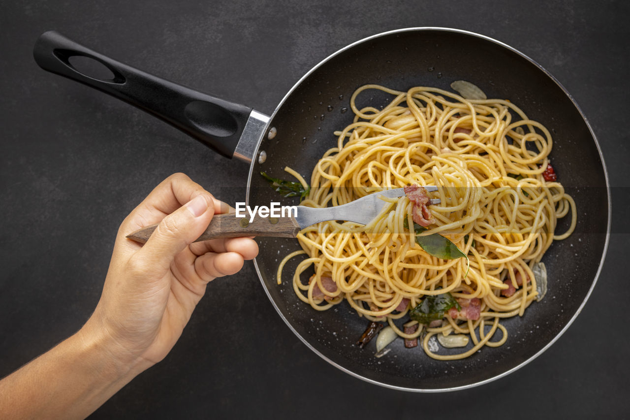 Hand holding fork get spaghetti pasta with dried chilli, garlic, sweet basil and bacon in black pan