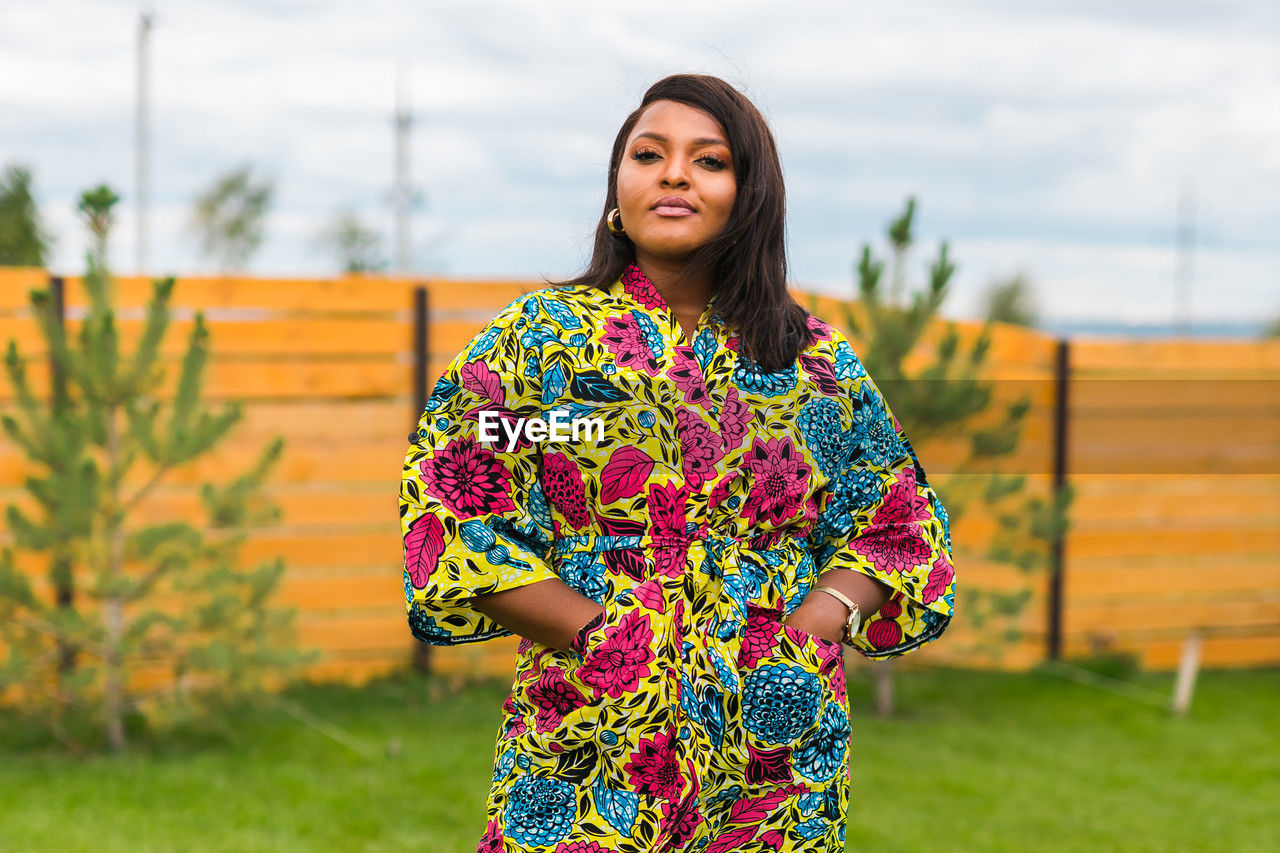 one person, yellow, women, adult, spring, smiling, standing, young adult, happiness, portrait, nature, dress, clothing, lifestyles, emotion, multi colored, flower, hairstyle, sky, female, grass, front view, cheerful, pattern, outdoors, focus on foreground, floral pattern, plant, fashion, photo shoot, looking at camera, architecture, waist up, leisure activity, day, person, casual clothing, copy space, brown hair, long hair, three quarter length, green, human face, landscape, relaxation, enjoyment
