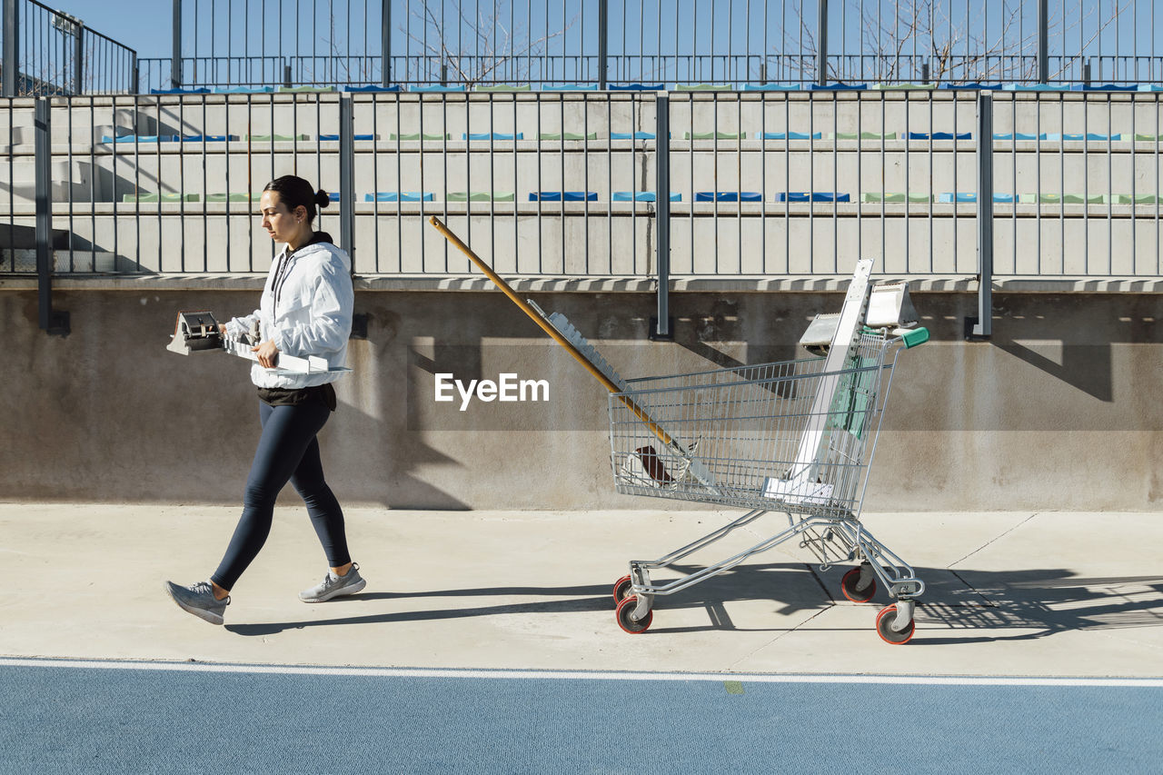 Side view of young female athlete carrying starting blocks while walking near cart with gear before track and field training on sunny day on stadium