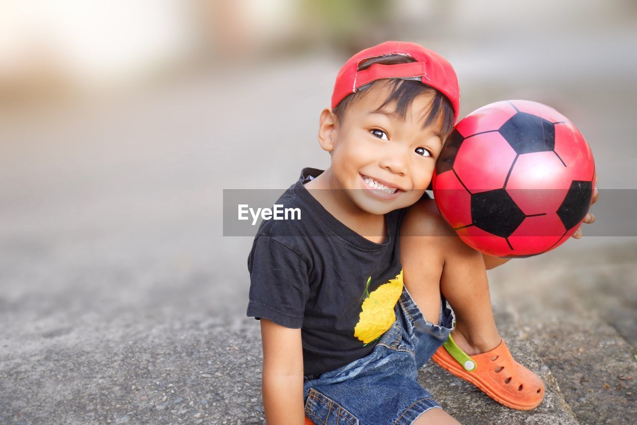 Portrait of cheerful boy sitting with ball on rock