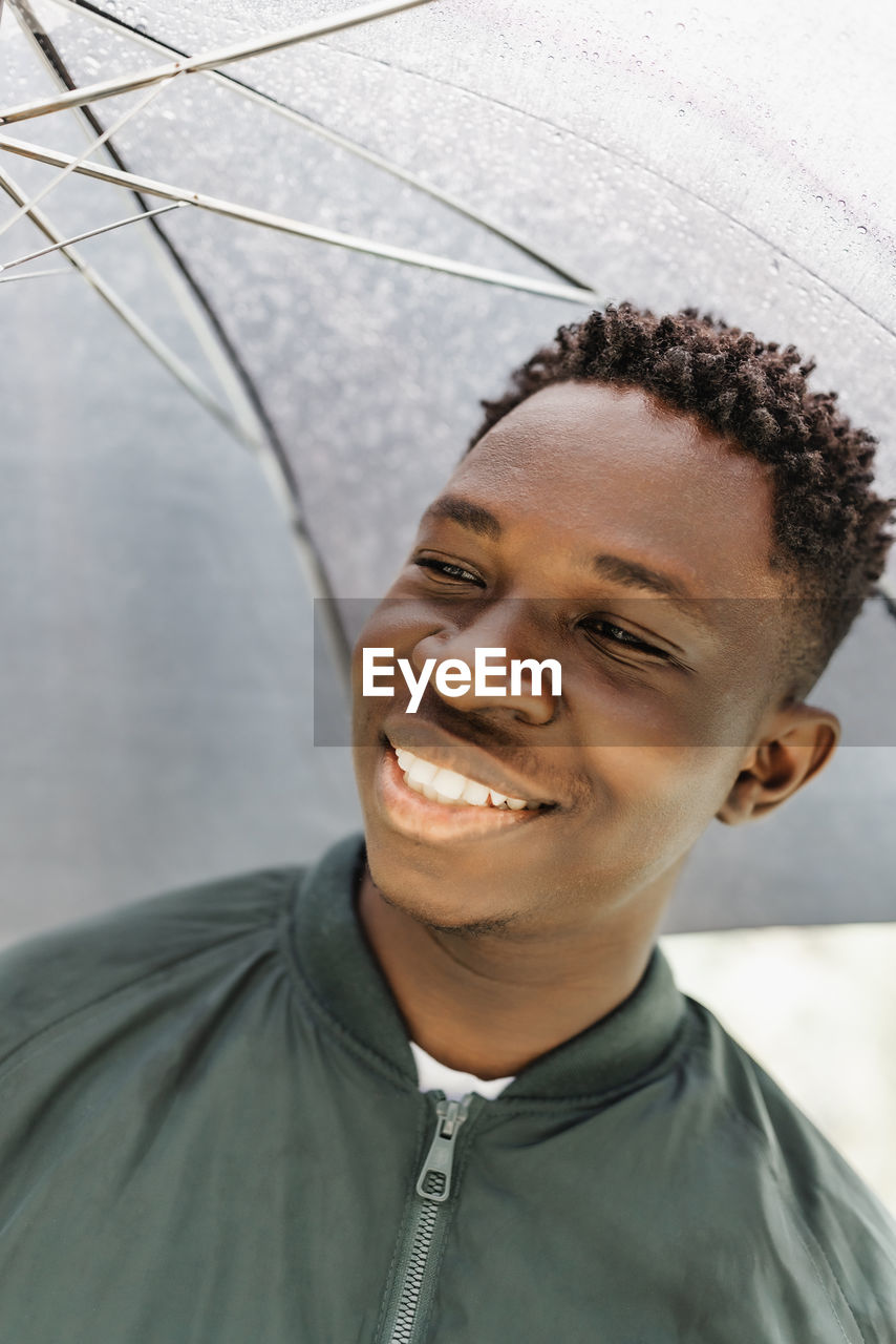 Young african american man under black umbrella in rain, smiling. fall or spring weather