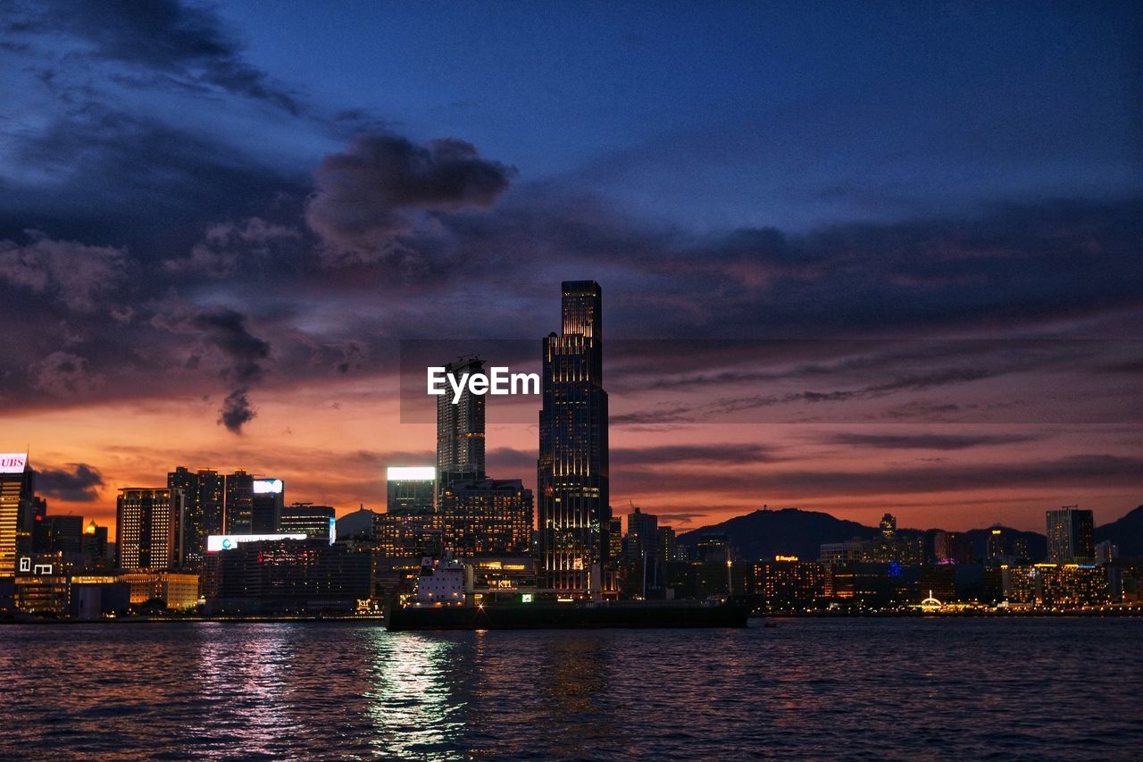 Illuminated modern buildings by river against sky at sunset