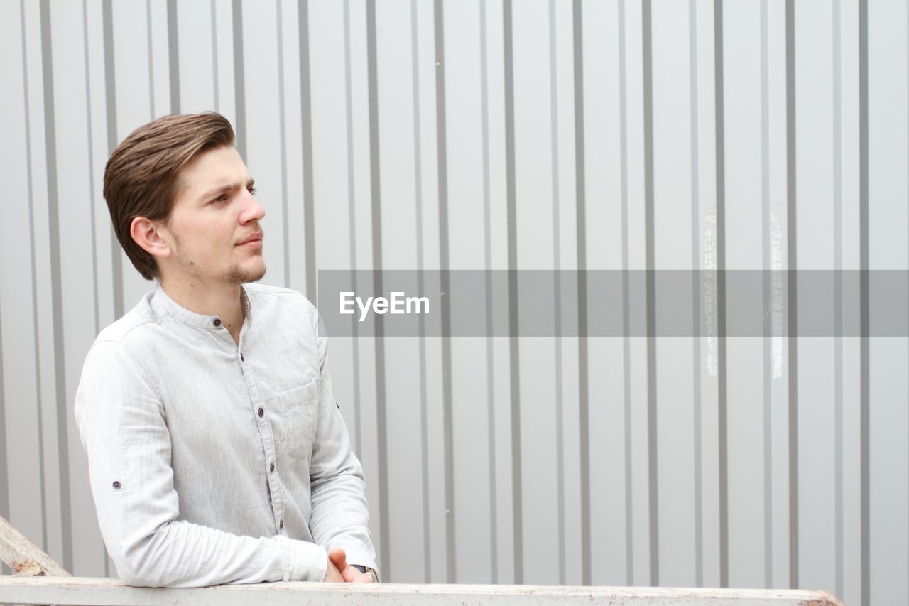 Young man looking away while standing against wall