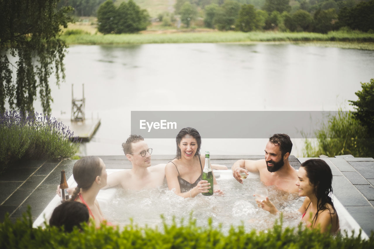 Cheerful male and female friends enjoying drinks in hot tub against lake during weekend getaway