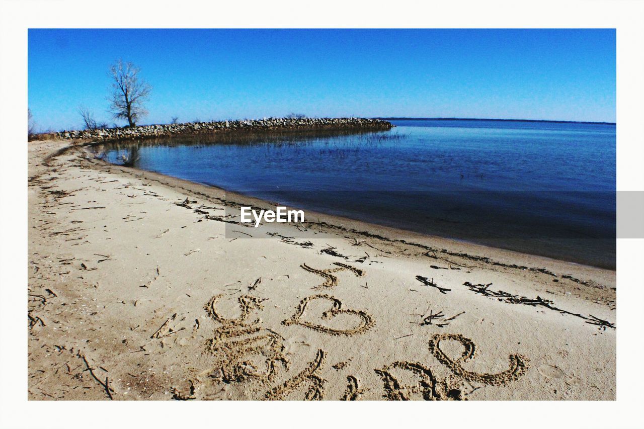 View of text with heart shape on sand against clear blue sky at beach