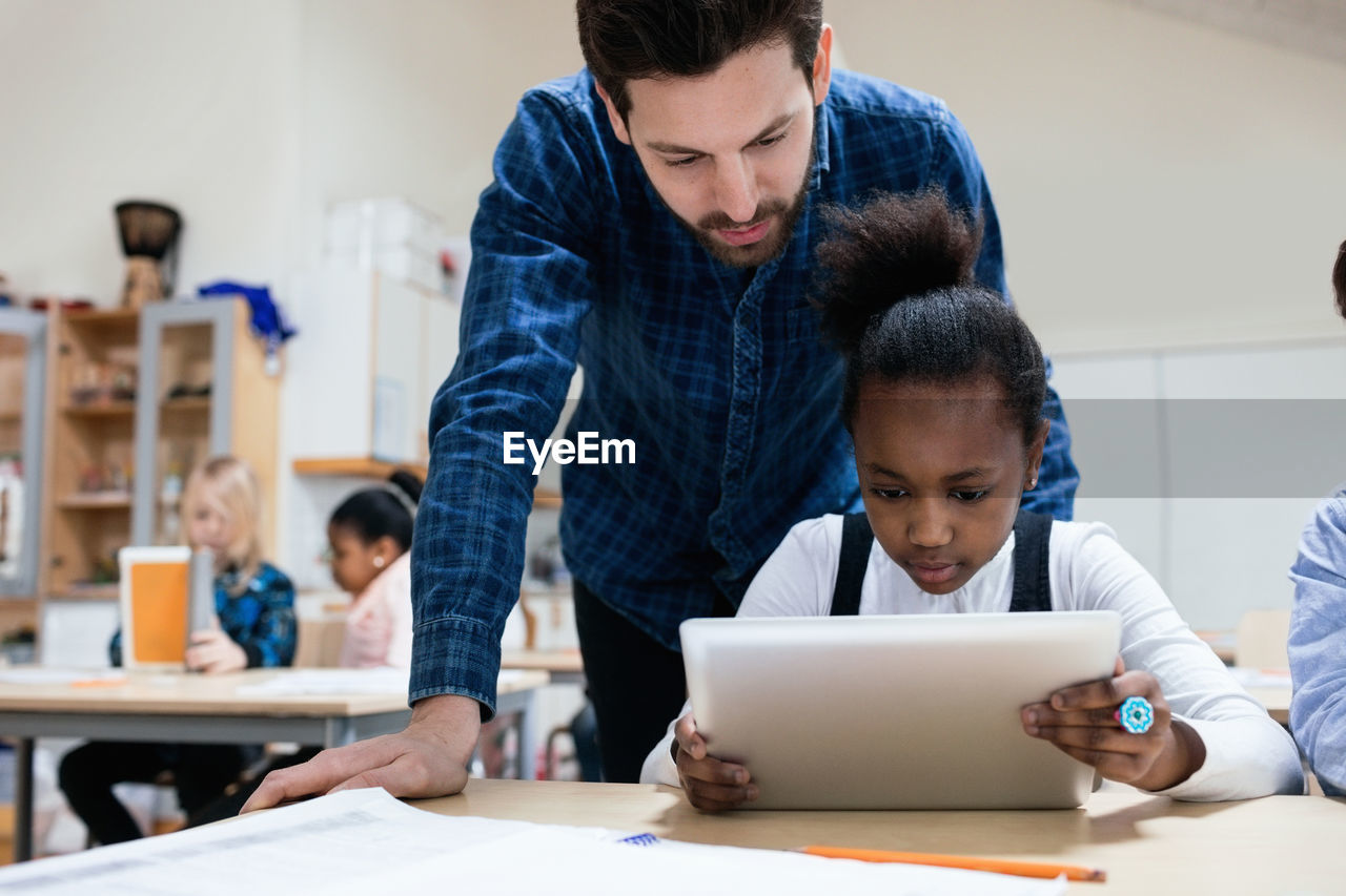 Teacher looking at girl using digital tablet in classroom