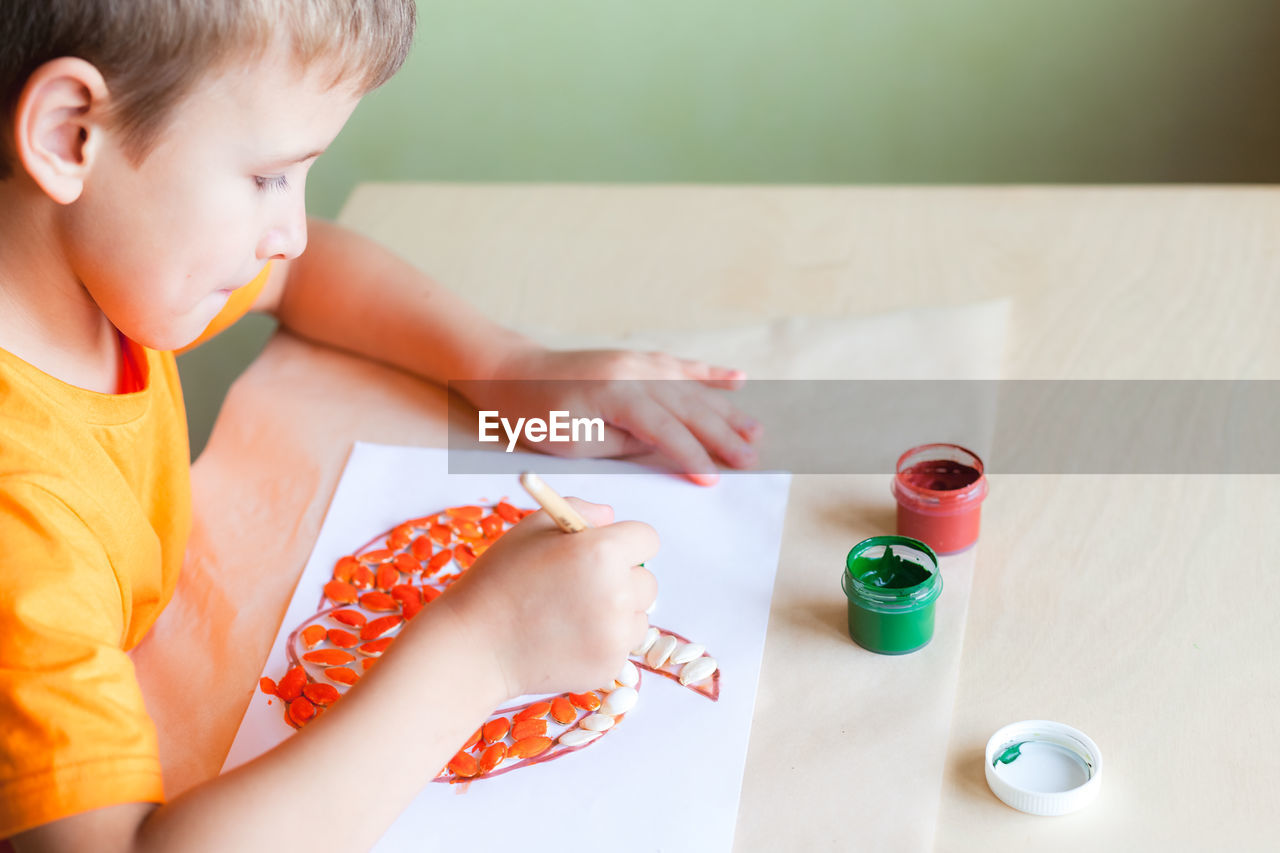 School boy making pumpkin from seeds on white paper, halloween diy concept. step by step instruction