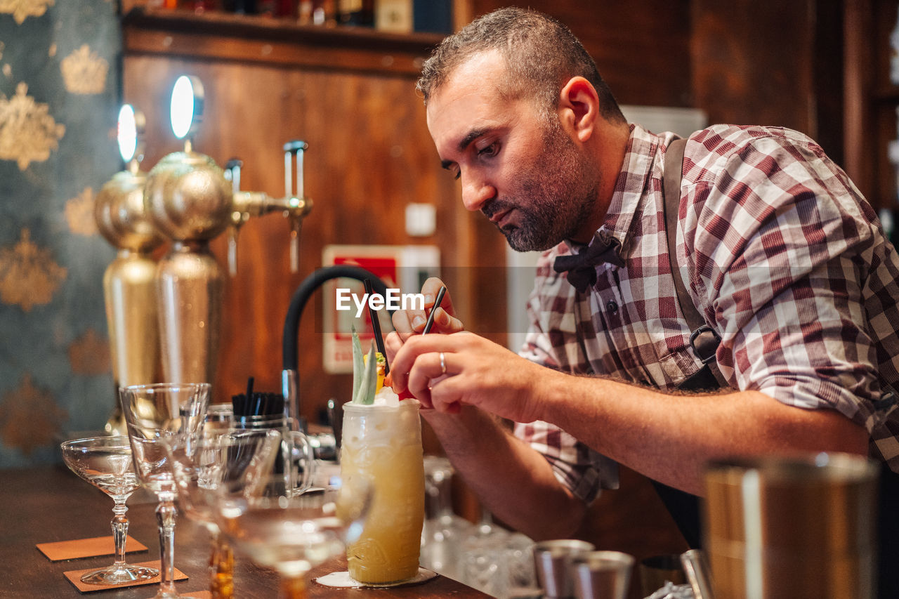 Concentrated mature barman in checkered shirt putting pieces of fruit and ice on top of tropical cocktail during work in bar