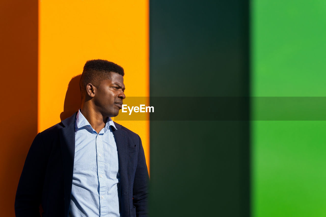 African american male in formal wear frowning face from sunshine while leaning on bright wall and looking away