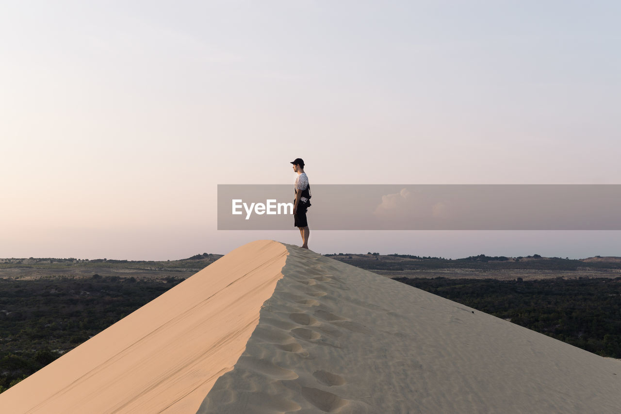Side view of man standing on sand dune against sky during sunset