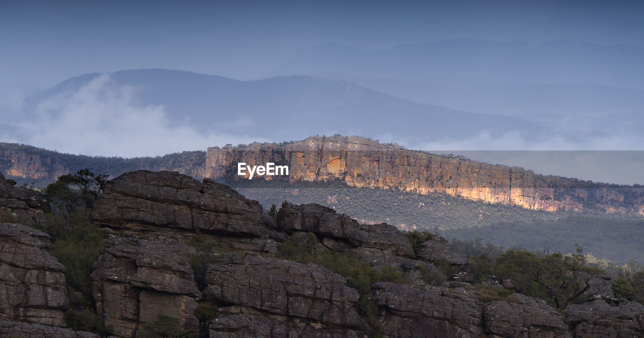 Scenic view of sunlit rocky mountains against sky with low wispy cloud