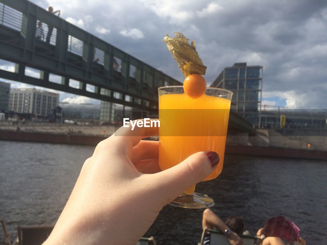 CLOSE-UP OF HAND HOLDING DRINK AGAINST RIVER