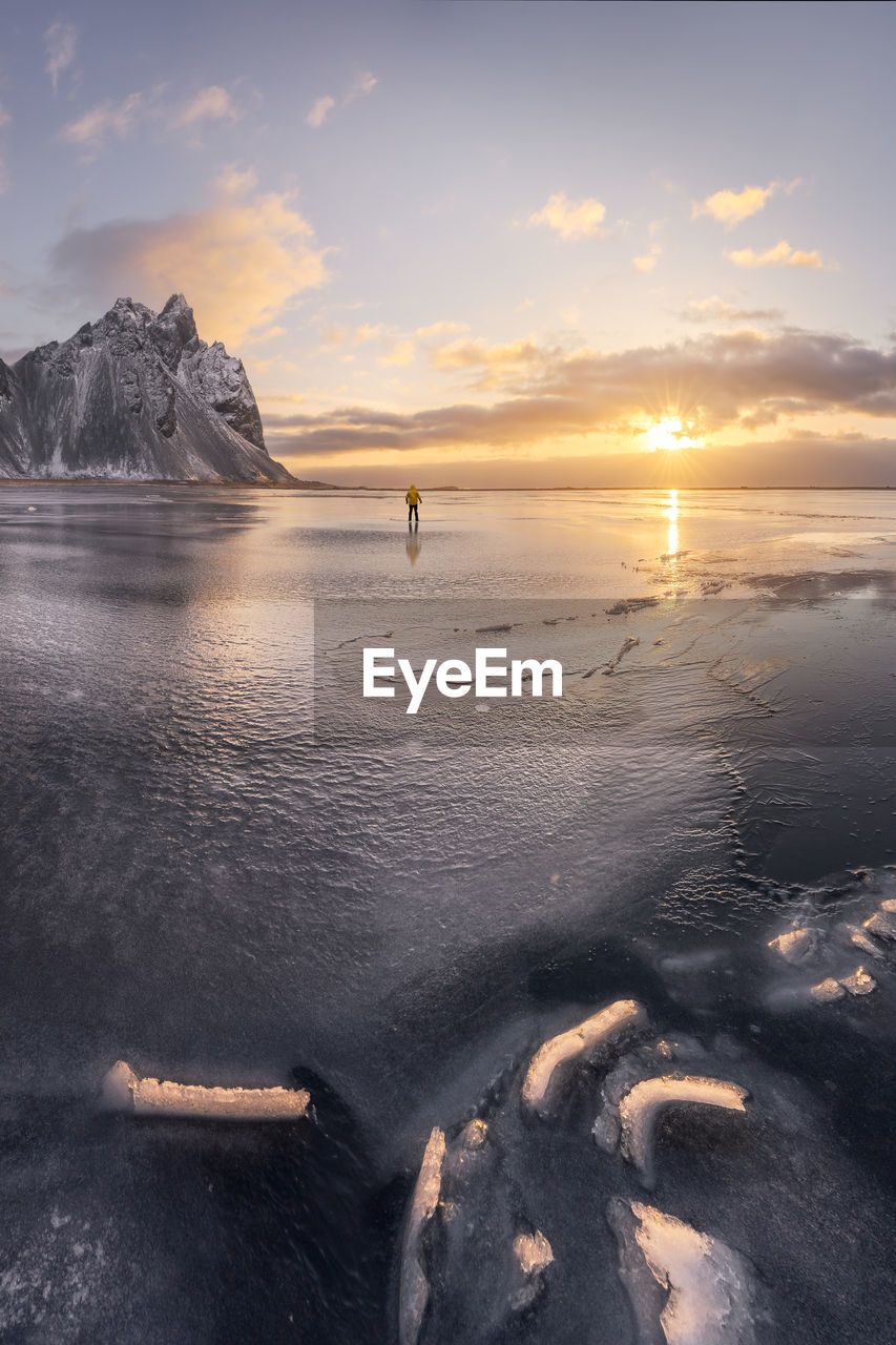 Anonymous tourist standing on wet shore near calm sea and rough mountains during majestic colorful sunset