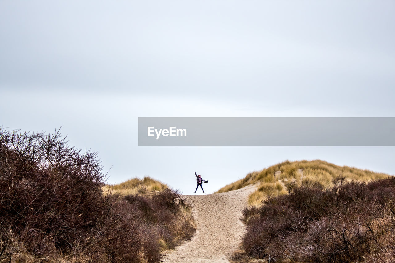 Woman jumping on dirt road amidst bush at hill against sky