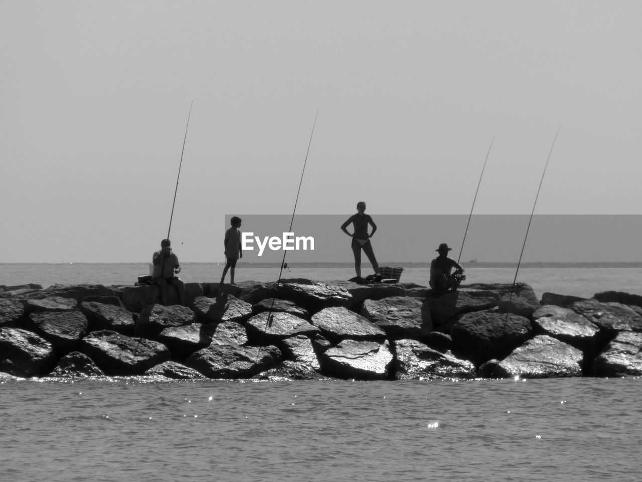 People with fishing rods standing on rocks amidst sea against clear sky during sunny day