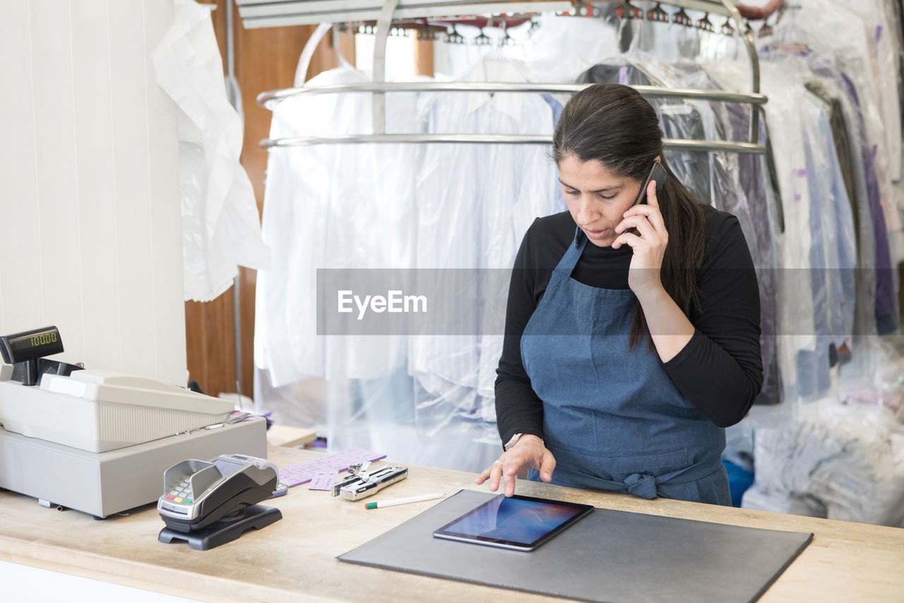 Mature female dry cleaner using digital tablet while talking on smart phone at laundromat