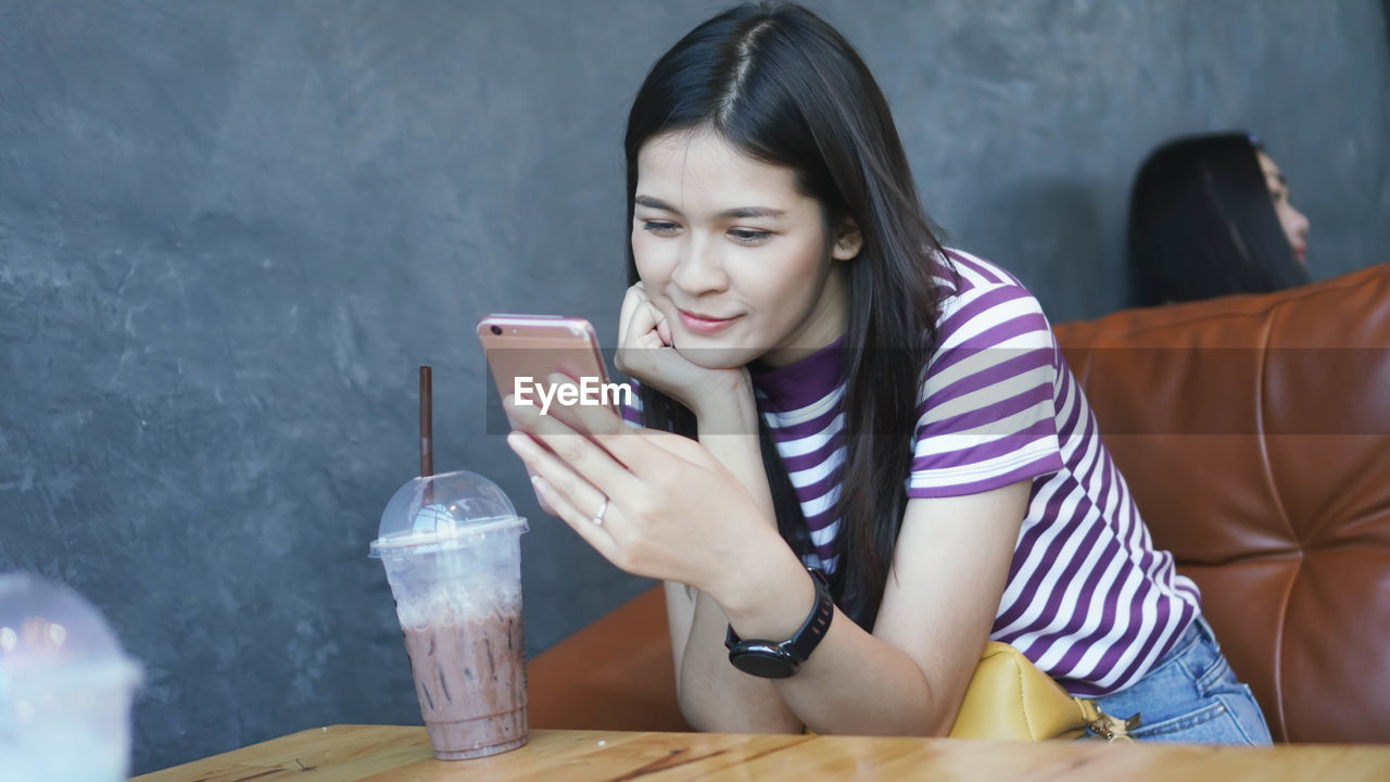 Smiling young woman using smart phone sitting at table with coffee in cafe 