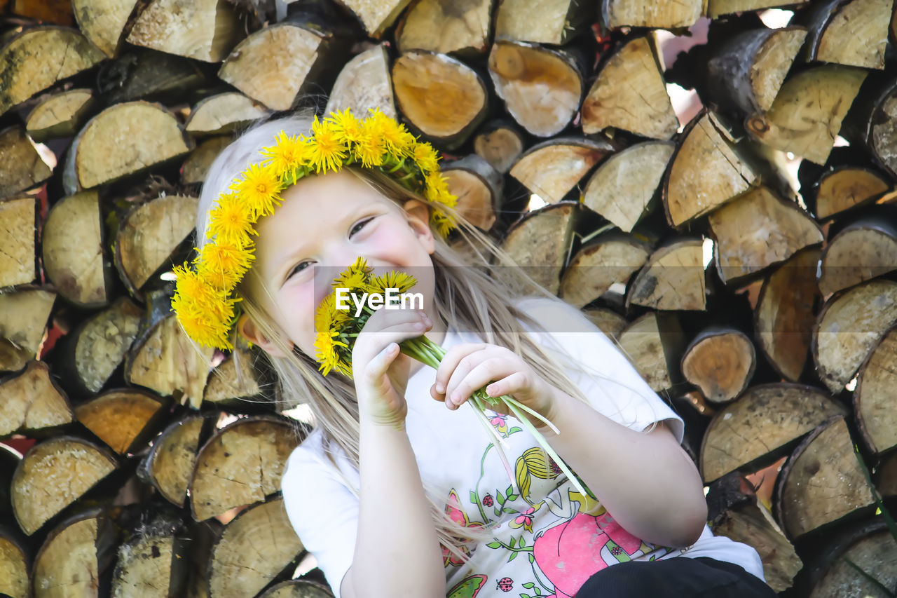 A little girl with a wreath on her head sitting near the firewood stack outdoors.