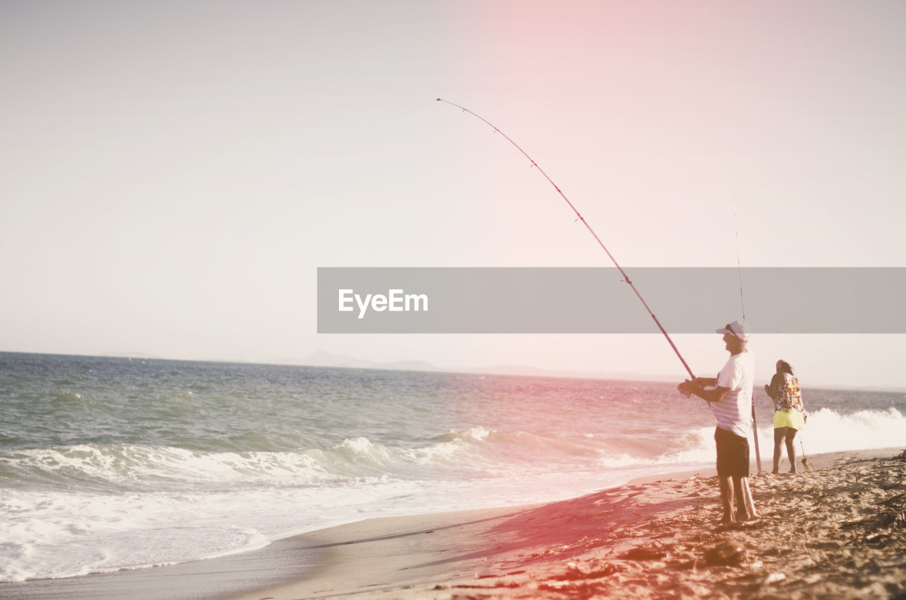 Rear view of men fishing at beach against clear sky