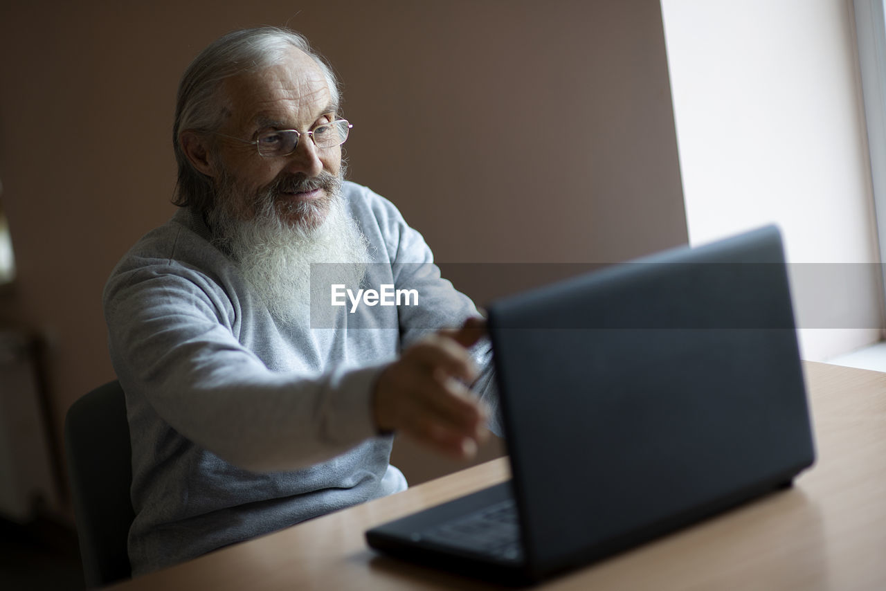 Smiling pensive old man with eyeglasses sitting at the desk and looking at laptop