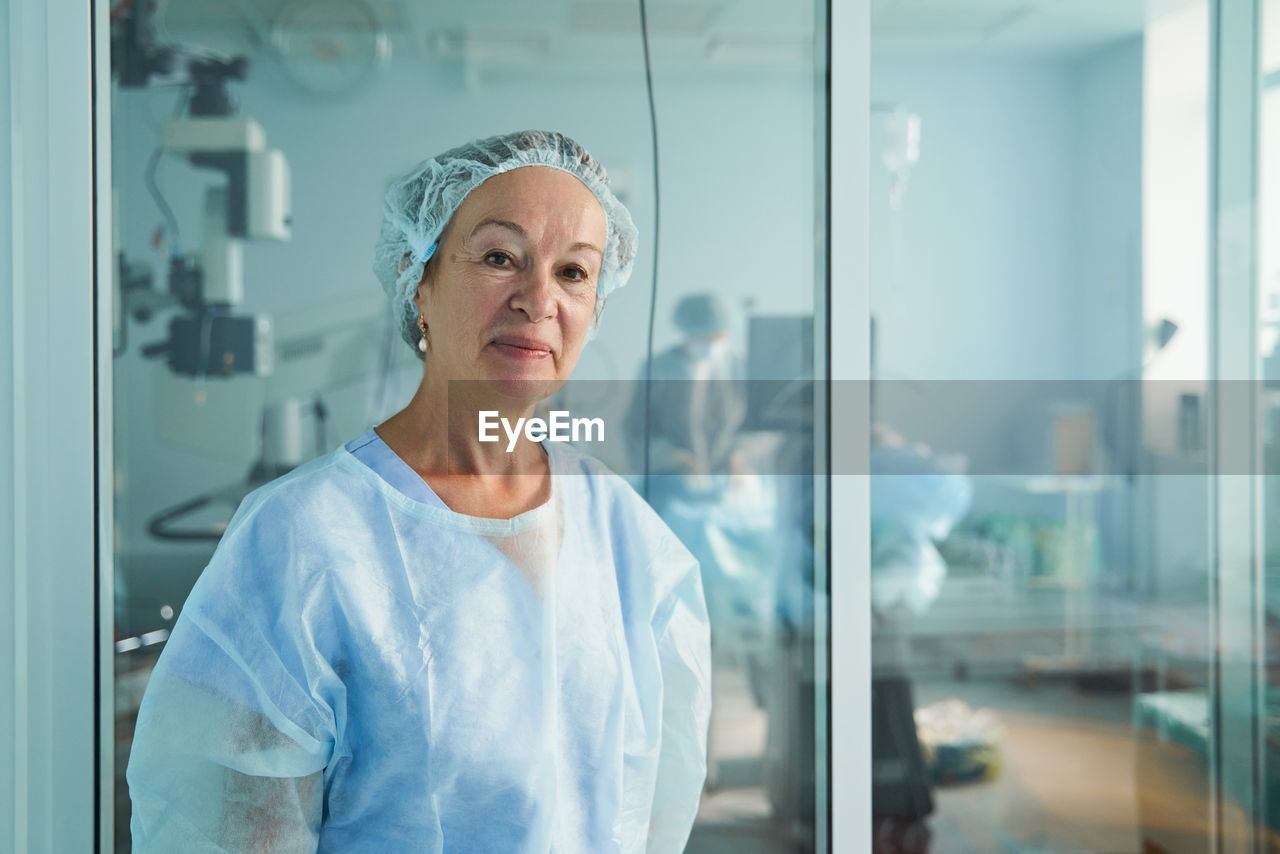 Friendly middle aged female doctor in surgical uniform looking at camera against unrecognizable colleague in hospital