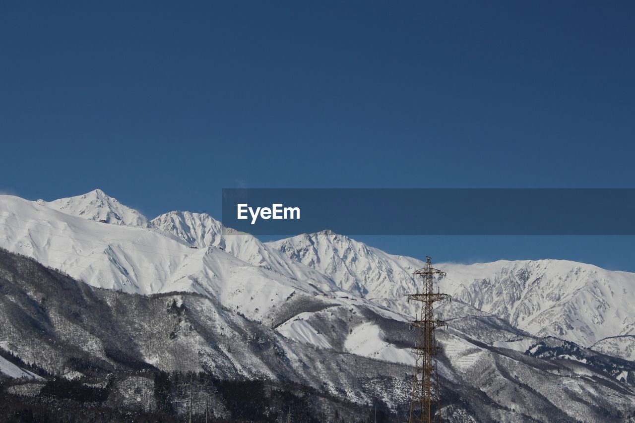 Low angle view of electricity pylon by snow covered mountains against clear blue sky