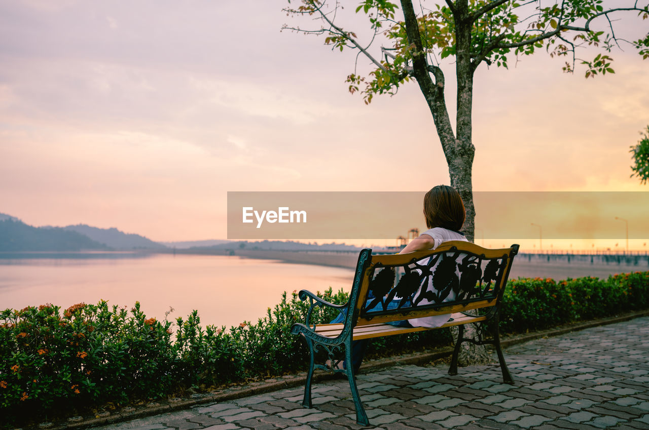 Rear view of mature woman looking at lake while sitting on bench against sky during sunset