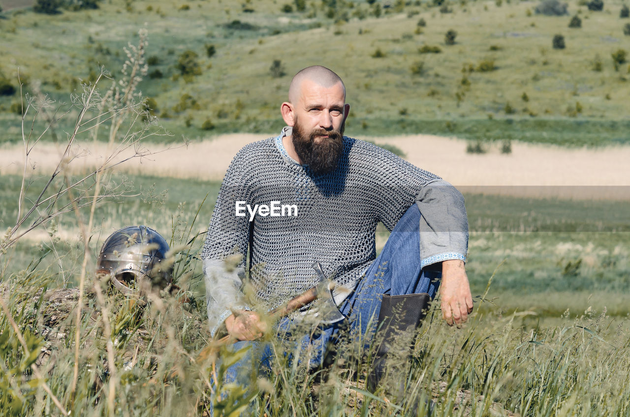 Viking. bald bearded man in metal chain mail over linen shirt sits on hill, holding ax. helmet lies