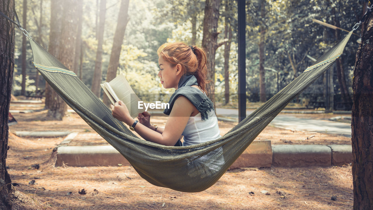 Side view of woman reading book while sitting on hammock in forest