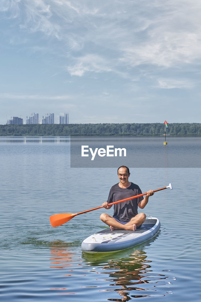 Asian older man sitting on sup board, rowing with oar on calm lake. good moments concept.