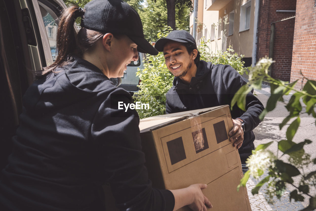 Smiling delivery colleagues holding cardboard box near truck