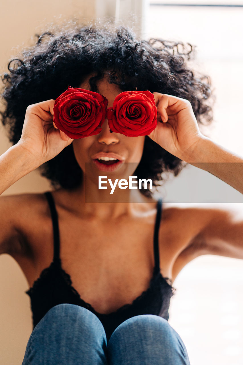 High angle of content african american female with afro hairstyle covering eyes with buds of red rose flowers at home