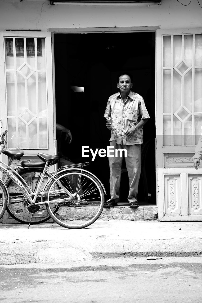 FULL LENGTH PORTRAIT OF MAN WITH BICYCLE STANDING AGAINST WALL