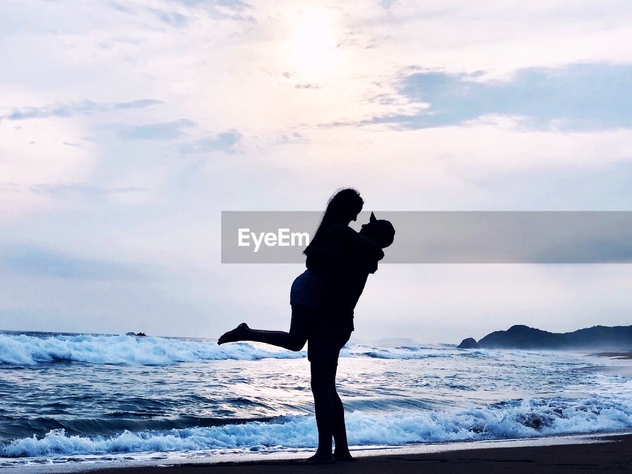 Side view of silhouette boyfriend carrying girlfriend on shore at beach against sky