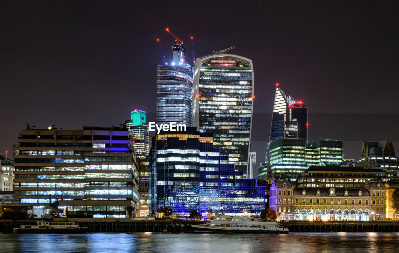 Illuminated buildings by river against sky at night