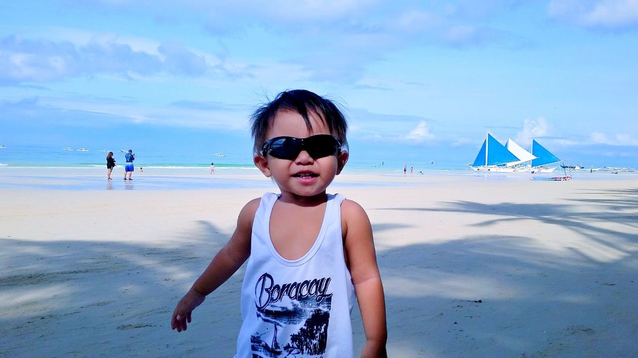 Portrait of baby boy in sunglasses standing at beach against sky