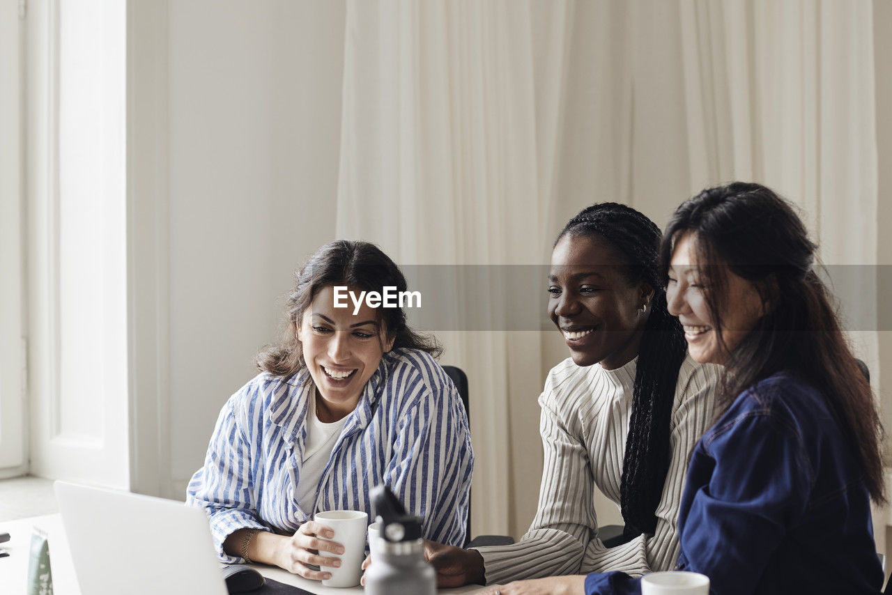 Happy female business professionals watching laptop while sitting at desk in coworking office