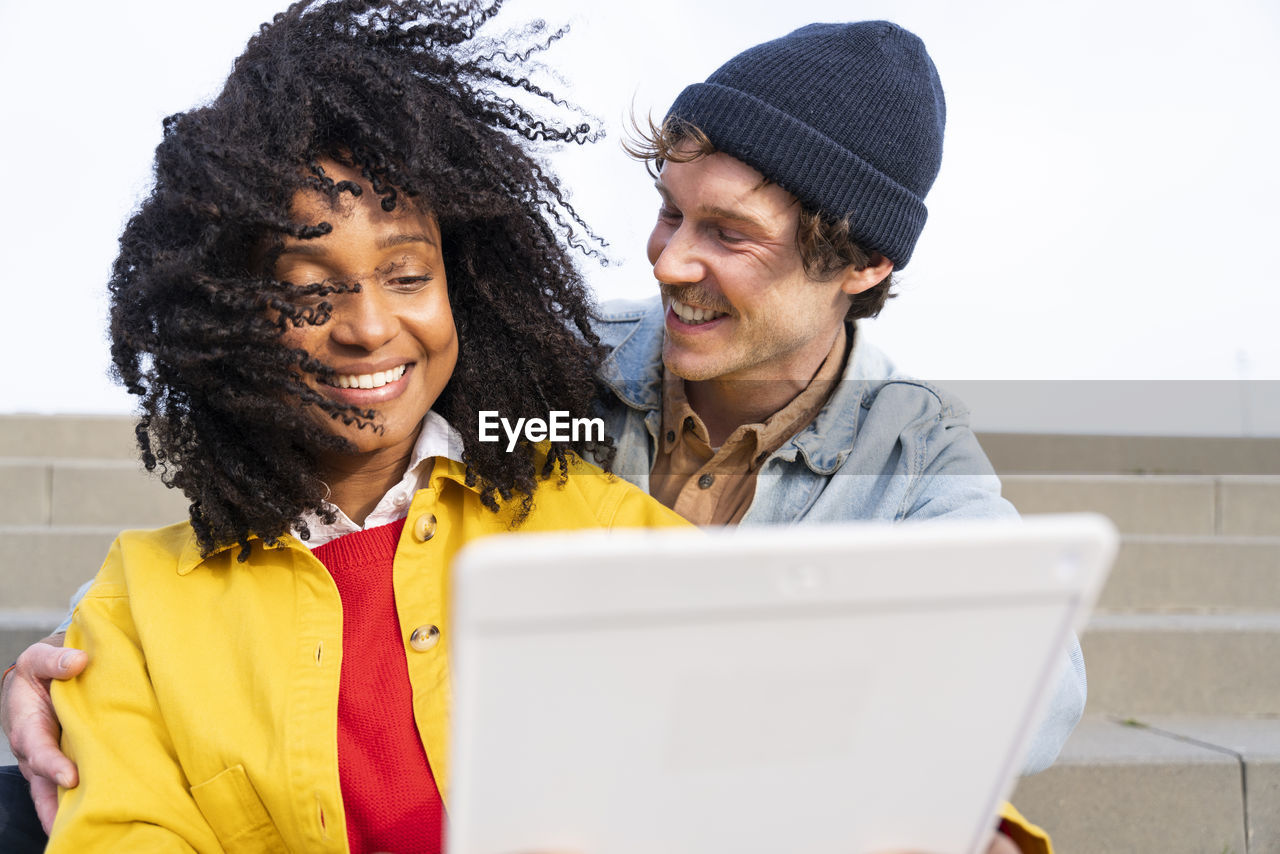 Happy man looking at girlfriend with tousled hair using tablet pc